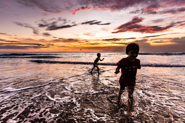 Two little boys having fun while running through water on the beach at sunset. Copy space.