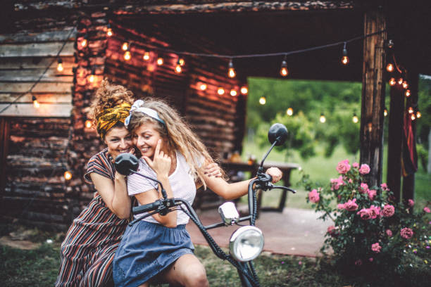 Two young friends ride a scooter on a scenic rural road. The driver wears a white polo shirt and a full-face helmet with goggles, while the passenger, wearing a black hijab and patterned blouse, smiles warmly. The lush greenery in the background enhances the joyful and adventurous atmosphere.