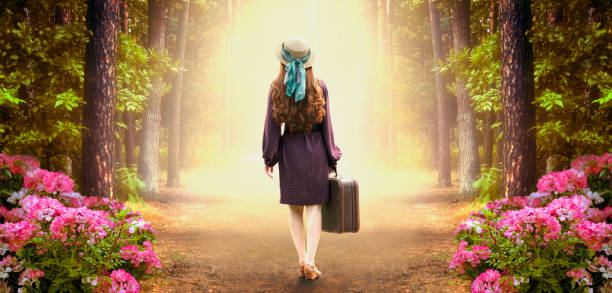 Young redhead woman in polka dot dress and hat with suitcase in retro style walking along a summer pine forest path to mystical glow. Travel to fairytale concept. Young redhead woman in polka dot dress and hat with suitcase in retro style walking along a summer pine forest path to mystical glow. Relaxation and vacation in woodland. Travel to fairytale concept. single lane road footpath dirt road panoramic stock pictures, royalty-free photos & images