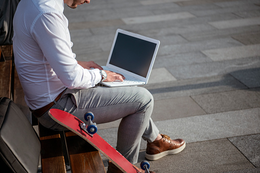freelancer working with laptop and smartphone while sitting on bench with longboard
