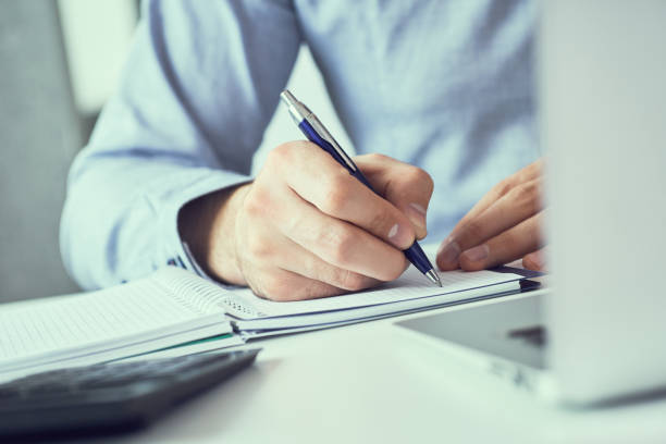istock Businessman hand writing note on a notebook. Business man working at office desk. Close up of empty notebook on a blackboard with office supplies. 1154808599