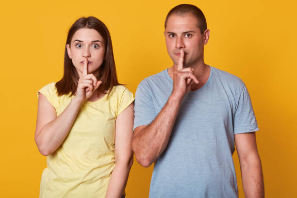 istock Serious young couple standing close near each other, touching their lips with forefingers, making gestures, keeping in silent, looking directly at camera, wearing casual clothes. Family concept. 1154809821