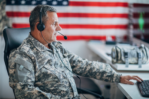 One senior soldier in uniform using headphones with microphone and computer at the office with American flag on the wall.