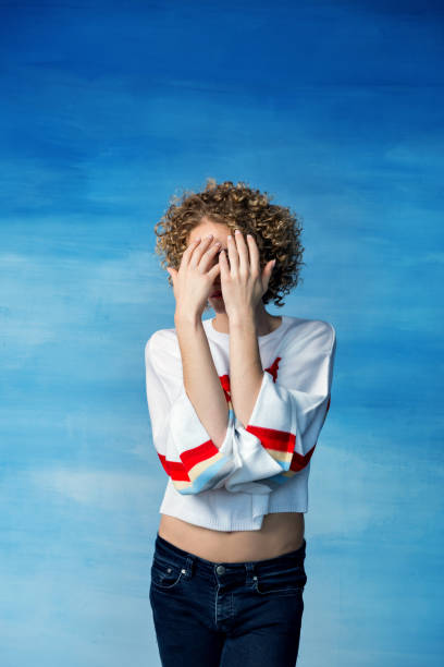 A young transgender guy in a white sweater with a rainbow symbol covered his face with hands on a blue background.