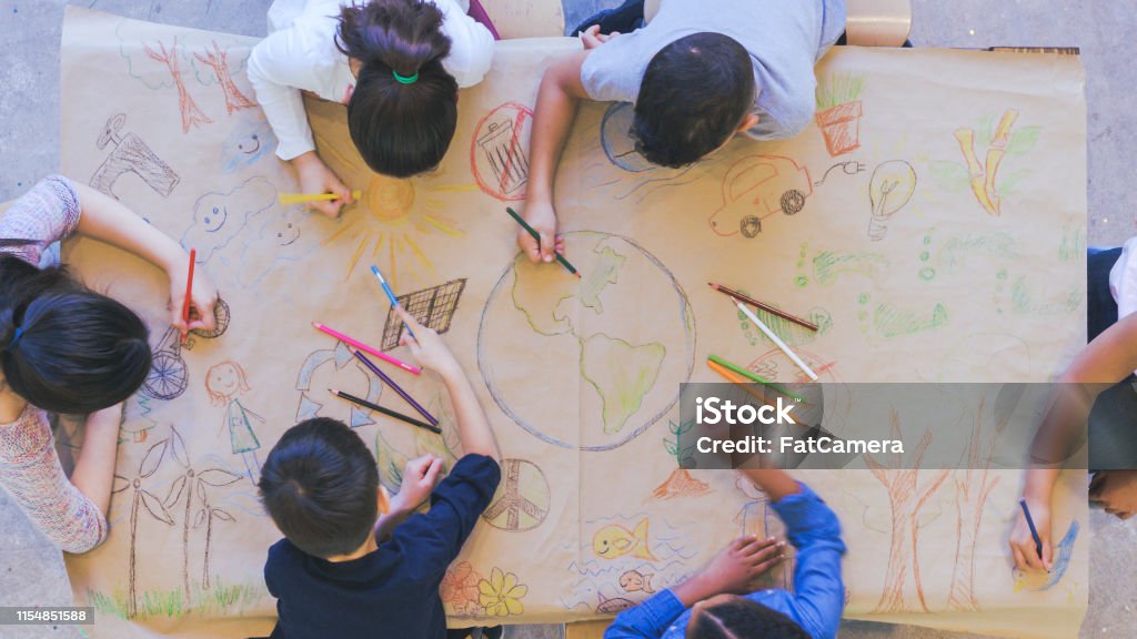 Group of children color environmentally conscious mural Aerial overhead view of a multi-ethnic group of elementary age children drawing. They are seated around a table. The kids are using colored pencils to make a mural. The have colored a world map, objects found in nature, and symbols of environmental conservation. Child Stock Photo