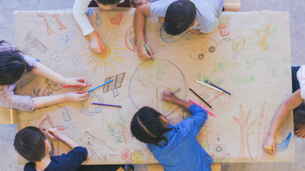 Group of children color environmentally conscious mural Aerial overhead view of a multi-ethnic group of elementary age children drawing. They are seated around a table. The kids are using colored pencils to make a mural. The have colored a world map, objects found in nature, and symbols of environmental conservation. social development kids stock pictures, royalty-free photos & images