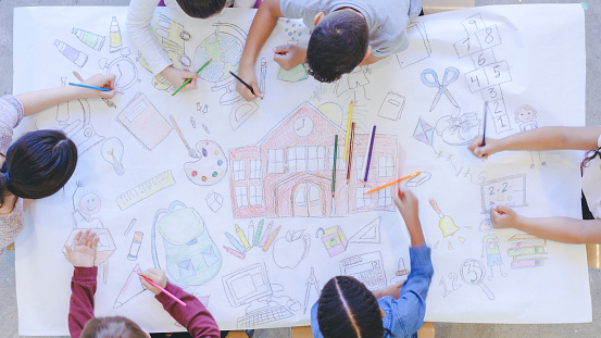 Aerial overhead view of multi-ethnic group of elementary age children coloring. The kids are seated around a table. They are creating a mural with colored pencils on paper. The children have colored a school and various school supplies.