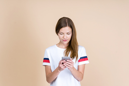 Front view of adult girl holding smartphone in hands, smiling and standing isolated on pastel beige background