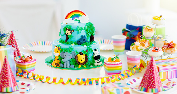 Cake for kids birthday celebration. Jungle animals theme children party. Decorated room for boy or girl kid birthday. Table setting with presents, gift boxes, confetti and sweets. Pastry for child