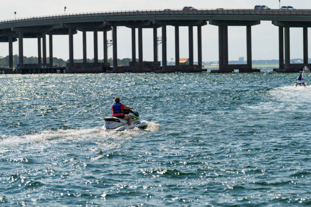 Two women skiing on jet ski outdoors at Gulf of Mexico ocean with view on US route 98 road bridge in Harborwalk Village, Florida Panhandle Destin, USA - April 24, 2018: Two women skiing on jet ski outdoors at Gulf of Mexico ocean with view on US route 98 road bridge in Harborwalk Village, Florida Panhandle harborwalk stock pictures, royalty-free photos & images