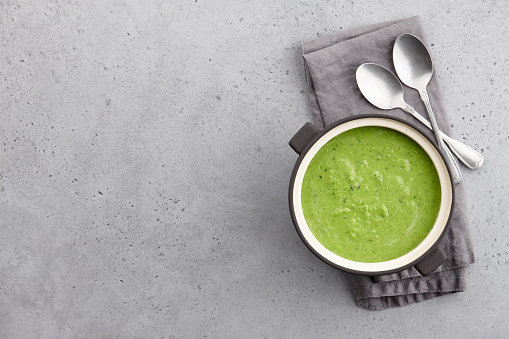 Fresh green pea soup served in bowl with spoons and napkin on gray concrete background. Top view, copy space.