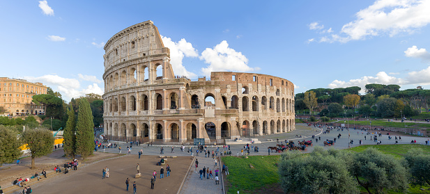 Panorama of the Coliseum in Rome is captured on a sunny autumn day. Around the majestic old building, there are many tourists. The sky above the Coliseum is blue.