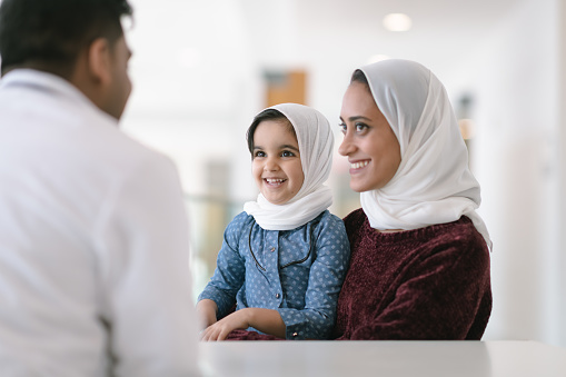 A young muslim mother wearing a hijab takes her toddler daughter to visit the pediatrician. The girl is also wearing a hijab and is sitting on her mother's lap. They are seated at a table and a male doctor is seated across from them. The mother and child are smiling at the doctor.