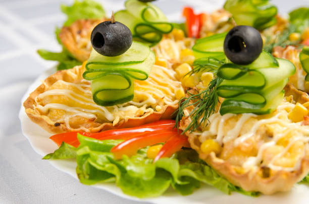 white dish with several tartlets with chicken, ham, cucumbers, lettuce and olive on white tablecloth.