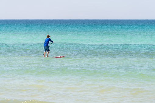 Santa Rosa Beach, USA - April 25, 2018: Man in hat shirt and shorts paddling on stand up board, surfboard in Gulf of Mexico water with waves, horizon in Pensacola Panhandle Florida