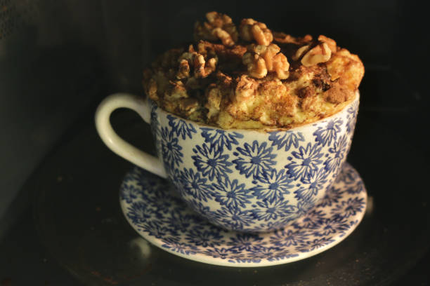 Image of homemade mug cake recipe of sponge after being cooked in kitchen microwave oven, topped with walnuts and brown sugar, carrot cake recipe made and cooking in mug / teacup on glass turntable, sweet pudding desert unhealthy snack after dinner treat