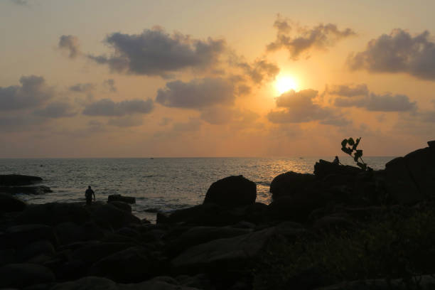 immagine del tramonto della spiaggia arancione che si riflette sul mare scintillante sull'acqua mentre il sole tramonta all'orizzonte, con sagome di rocce e persone / turisti che guardano il tramonto scattare foto delle vacanze con telecamere, alba / tramo - sunset vacations orange glowing foto e immagini stock