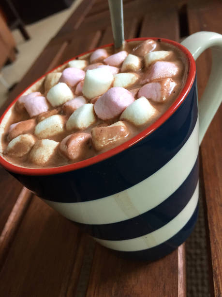 Stock photo of frothy hot chocolate drink in mug with mini marshmallows. The hot chocolate drink mug is placed on a wooden table as background. The hot chocolate is topped with delicious pink and white marshmallow sweets imagen de la bebida de chocolate caliente en la taza cubierto con leche, crema batida y mini malvaviscos veganos/vegetarianos, dulces de caramelo rosa y blanco fusión, deliciosa receta de chocolate caliente gruesa en mesa de café de salón de madera para
