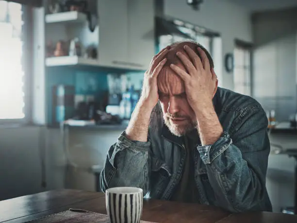 A mature man sits at a table at home, his head in his hands, looking thoroughly depressed and miserable.