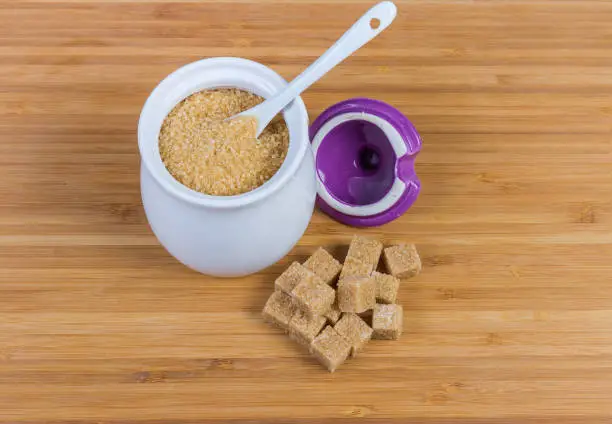 Natural unrefined brown granulated sugar in white sugar-bowl with spoon and cubes of brown sugar scattered beside to her on a wooden surface