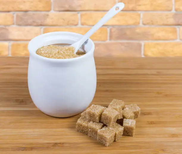 Natural unrefined brown granulated sugar in white sugar-bowl with spoon and cubes of brown sugar scattered beside to her on a wooden surface