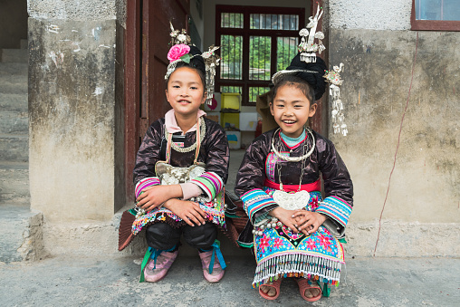 Two chinese dong people school girls sitting together in front of the entrance to their school classroom in traditional clothing in the small Dong Village Huang Gang - Huanggang, Shuangjiang, Liping, Guizhou, China.