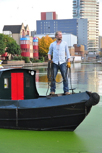 Regent's Canal in London wends its way through large swathes of the northern edge of the city centre. It passes through residential and commercial areas as well as former industrial areas transitioning to other uses. Many colorful wooden houseboats are moored along Regent's Canal and the canal is also a busy thoroughfare for these and other boats. Regent's Canal opens up into many scenic vistas and is therefore popular amongst sightseeing pedestrians as well as cyclists. Shown here: a houseboat captain prepares to enter the lock which will enable the boat to enter a part of the canal with a different elevation.  Photos taken on 10/25 in London.