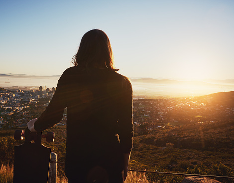 Half-length near silhouette of a young woman holding  a skateboard and looking out over a city at either sunrise or sunset.