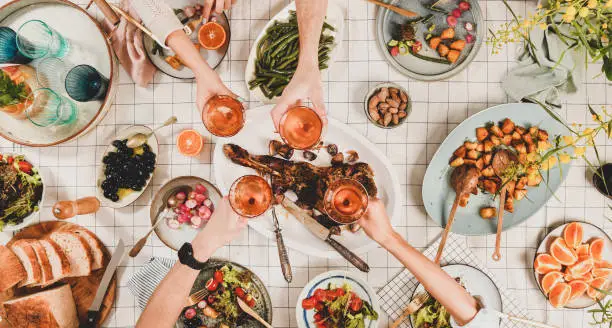 Family, friends gathering dinner. Flat-lay of peoples hands clinking with glasses of rose wine over table with roasted lamb shoulder, salad, vegetables, mimosa branch, top view. Celebration party