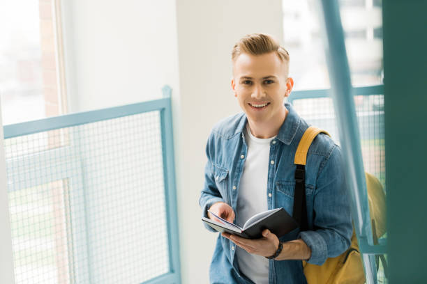 istock smiling student in denim shirt with yellow backpack holding notebook in university 1154970911