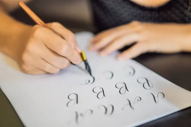 Calligrapher Young Woman writes phrase on white paper. Inscribing ornamental decorated letters. Calligraphy, graphic design, lettering, handwriting, creation concept.