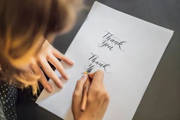 Thank you. Calligrapher Young Woman writes phrase on white paper. Inscribing ornamental decorated letters. Calligraphy, graphic design, lettering, handwriting, creation concept.