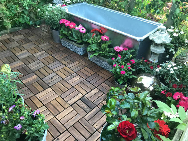 Stock photo of garden treehouse terrace platform balcony in summer with zinc metal trough pond water feature with solar fountain pump, goldfish fish, marginal plants, red miniature roses, pink gerbera flowers, teak decking tiles, solar powered lights, lighting imagen de jardín del árbol de la terraza de la plataforma balcón en verano con zinc metal levantado estanque de agua característica de la cubeta de ganado galvanizado listo para el agua, plantas, fuente, rosas en miniatura rojas, flores de gerbera rosa
