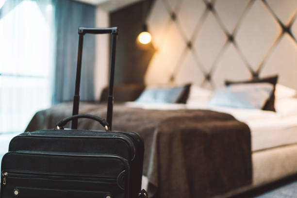 A black closed suitcase in a modern hotel room with lights on, brown, white and blue bedding and big windows.