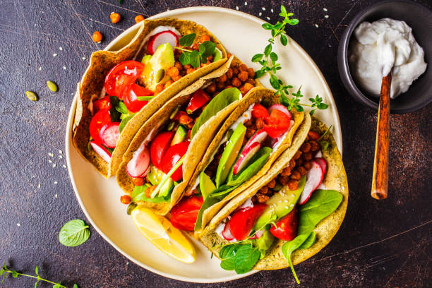 Vegan tacos with baked chickpeas, avocado, sauce and vegetables on dark background, top view.