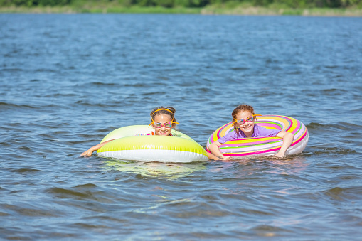 Two young girls (sisters) playing in innertube float toys on a lake in summer. The younger sister is on the right, with the older sister on the left. Both girls are wearing swim goggles and rash guard swim shirts. Taken on a beautiful summer day. In this image the girls are side by side and smiling at the camera.