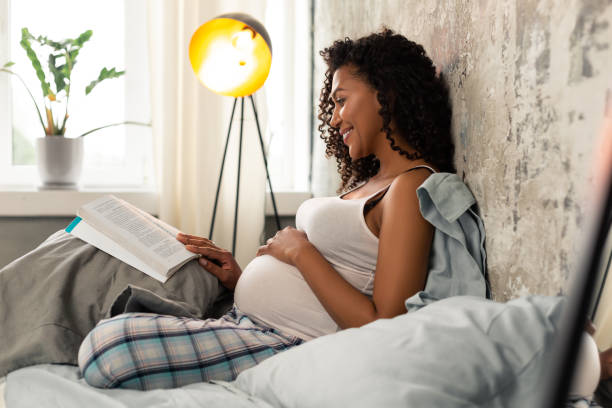 femme enceinte joyeuse lisant un livre dans son lit. - family reading african descent book photos et images de collection