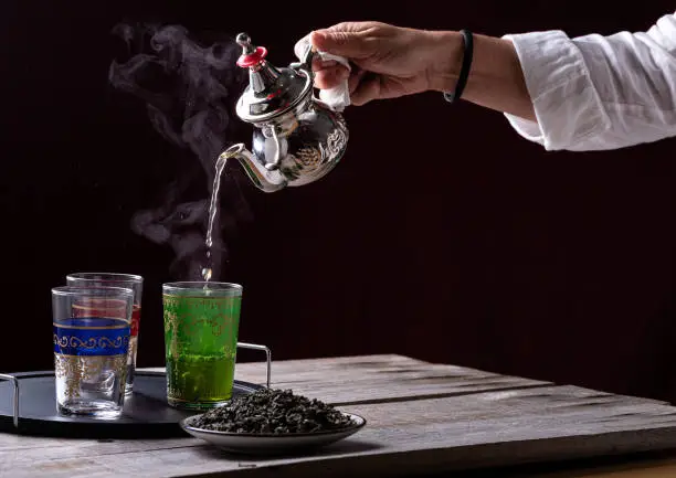 person serving tea in a silver teapot over glass cups