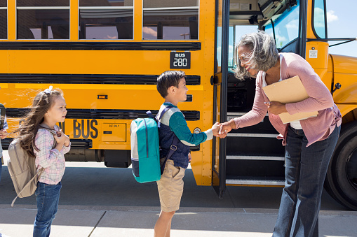 On his first day of school, a male child with a backpack on shakes the bus driver's hand outside the school bus.