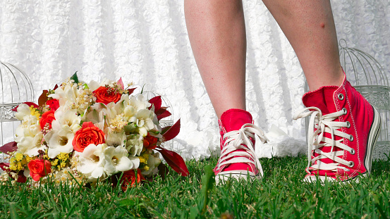 Legs of the modern bride in red sneakers and a bouquet on the background of green grass.