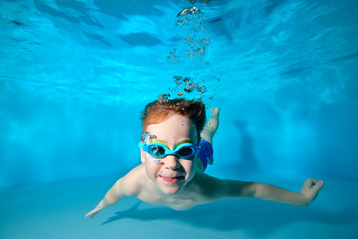 Happy smiling baby, boy, swimming underwater in swimming pool in swimming glasses and posing for camera on blue background. Portrait. Underwater photography. Vertical orientation.