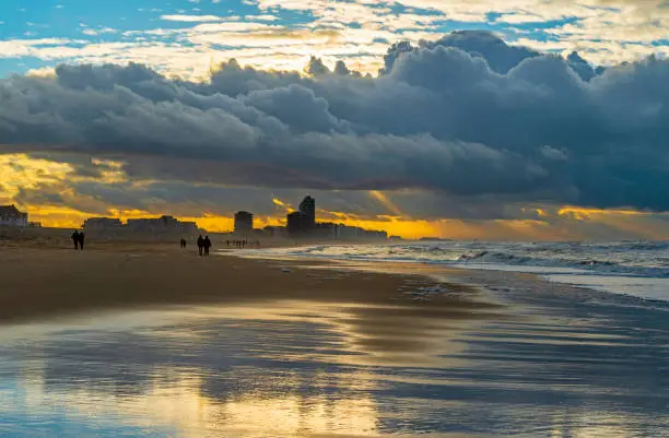 Sunbeam in the sky on Ostend beach during storm weather with its urban skyline and the silhouette of unrecognizable people walking along the shoreline of the North Sea with strong waves, West Flanders, Belgium.