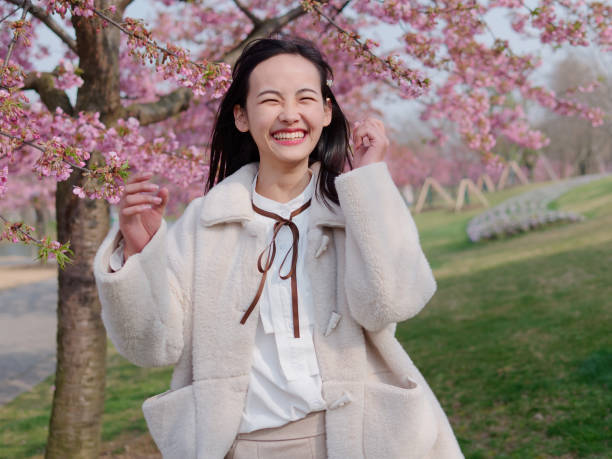 femme heureuse souriant et ayant l’amusement au jardin de cerise. portrait de printemps de la jeune belle fille en chemise blanche et manteau fourrure. fille chinoise riant et regardant l’appareil-photo avec la joie. - cherry blossom flower head spring flower photos et images de collection