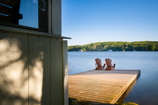 Adirondack chairs on a wooden dock on a calm lake in Muskoka, Ontario Canada. Cottages nestled between trees are visible across the water.
