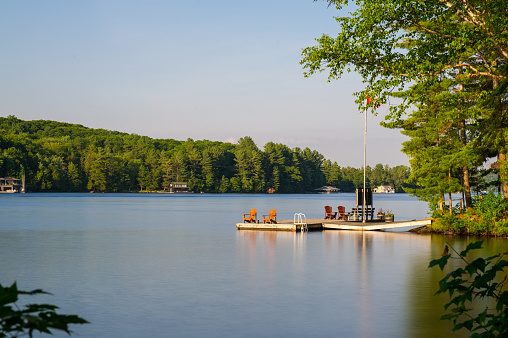 Muskoka chairs sitting on a wood dock facing a calm lake. Across the water is a cottage nestled among green trees. There is a sailing boat docked.
