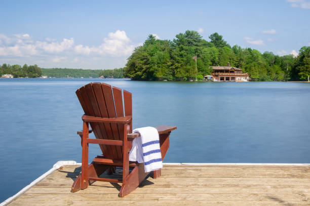 Adirondack chair on a wooden dock with a towel Adirondack chair on a wooden dock on a calm lake in Muskoka, Ontario Canada. A cottage nestled between trees is visible across the water. A white towel in folded on the arm chair. canada flag blue sky clouds stock pictures, royalty-free photos & images