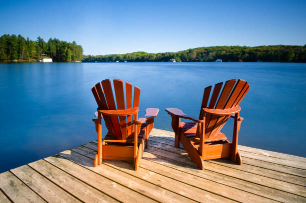 Adirondack chairs on a wooden pier Adirondack chairs on a wooden dock on a calm lake in Muskoka, Ontario Canada. Cottages nestled between trees are visible across the water. lake stock pictures, royalty-free photos & images
