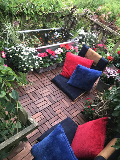 bild von gartenbaumterrasse plateauf-balkon im sommer mit roten und blauen kissen, zinkteich mit solarbrunnen pumpe, goldfische, randpflanzen, rote miniaturrosen, rosa gerbera blumen, teakdecking-fliesen, solar-lichter, beleuchtung - patio wood formal garden deck stock-fotos und bilder