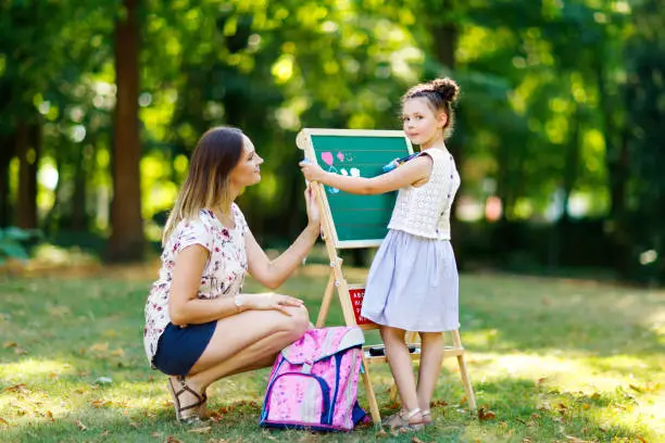 Happy little kid girl and mother by big chalk desk Preschool or schoolkid on first day of elementary class. Back to school concept. Healthy child and woman writing and painting outdoors.