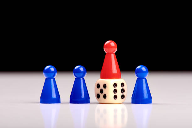 istock Three blue game pieces and between them one red piece stands on a dice as a winner or leader. Blurred black and white background 1155136389